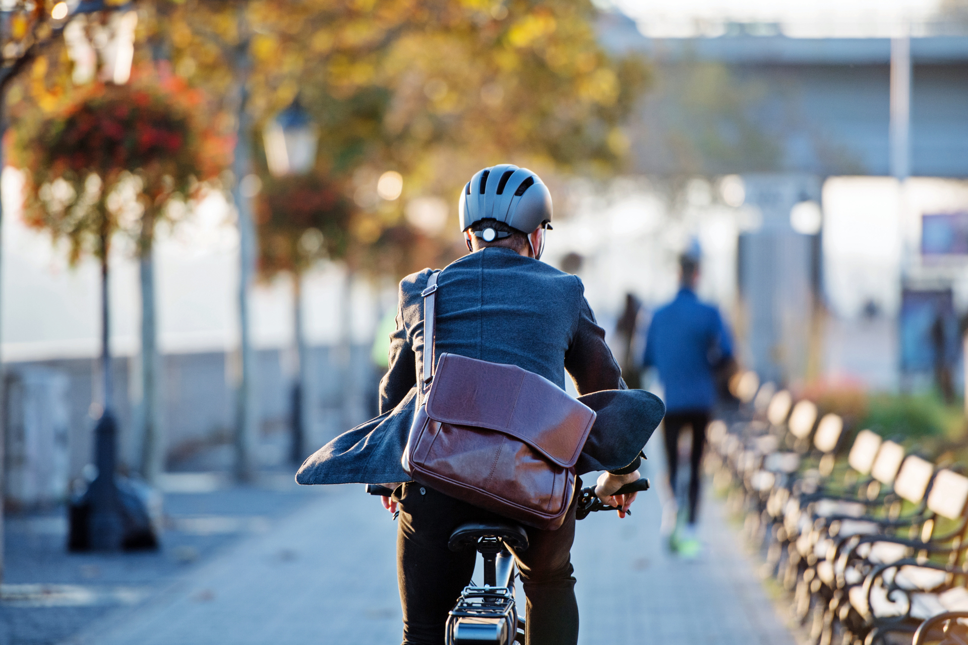 Ein Fahrradfahrer im Anzug mit Helm von hinten unterwegs an einer Promenade Ein Fahrradfahrer im Anzug mit Helm von hinten unterwegs an einer Promenade